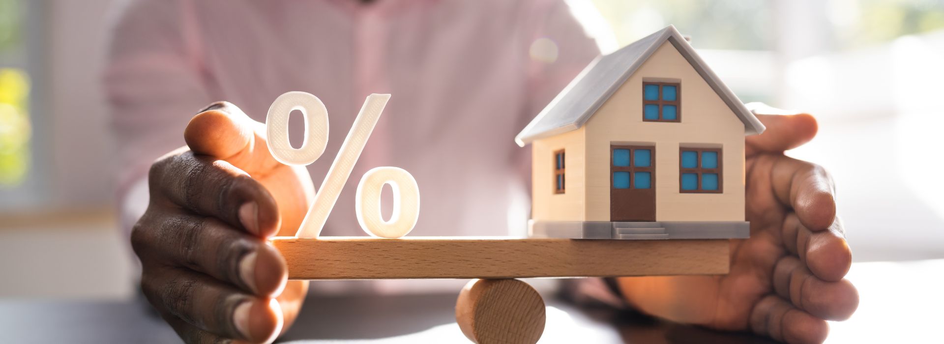 This image illustrates the concept of mortgage interest rates or home financing decisions. Two hands are positioned protectively around a wooden seesaw or balance scale. On one side of the balance sits a white percentage symbol (%), while on the other side is a small model house with cream-colored walls, a gray roof, blue windows, and a brown door. The wooden fulcrum beneath creates a teeter-totter effect, symbolizing the balance between interest rates and homeownership costs. The background is softly blurred with warm, natural lighting, giving the scene a professional yet approachable feel. This visual metaphor represents the careful consideration needed when weighing mortgage rates against property values in real estate and financial planning decisions.
