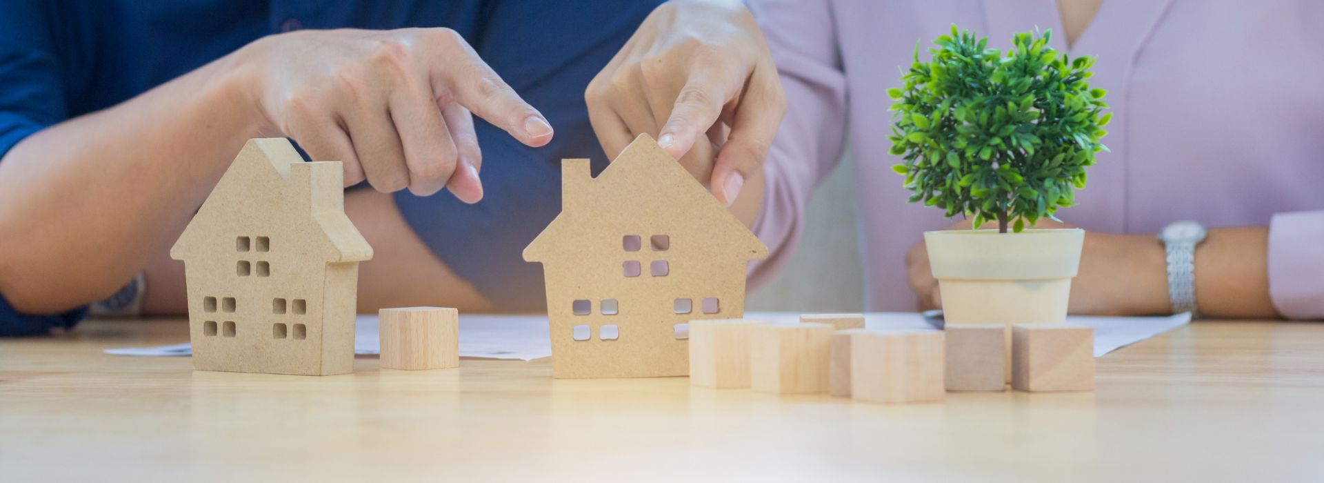 This image shows a real estate or property investment concept. Two people are seated at a light wooden table, with their hands positioned protectively over small wooden house models. The person on the left, wearing a dark blue shirt, has their hand covering a lighter-colored wooden house. The person on the right, in a light pink or lavender shirt and wearing a watch, is protecting a slightly larger wooden house model. Between and around the houses are several small wooden blocks or cubes arranged on the table surface. To the right side of the scene sits a small potted plant with bright green foliage in a cream-colored pot, adding a natural element to the workspace. The scene suggests themes of property protection, real estate planning, home insurance, or investment strategy discussions. The protective hand gestures over the model homes emphasize security and care in property-related decisions. Papers are visible on the table, indicating this might be during a consultation or planning meeting.