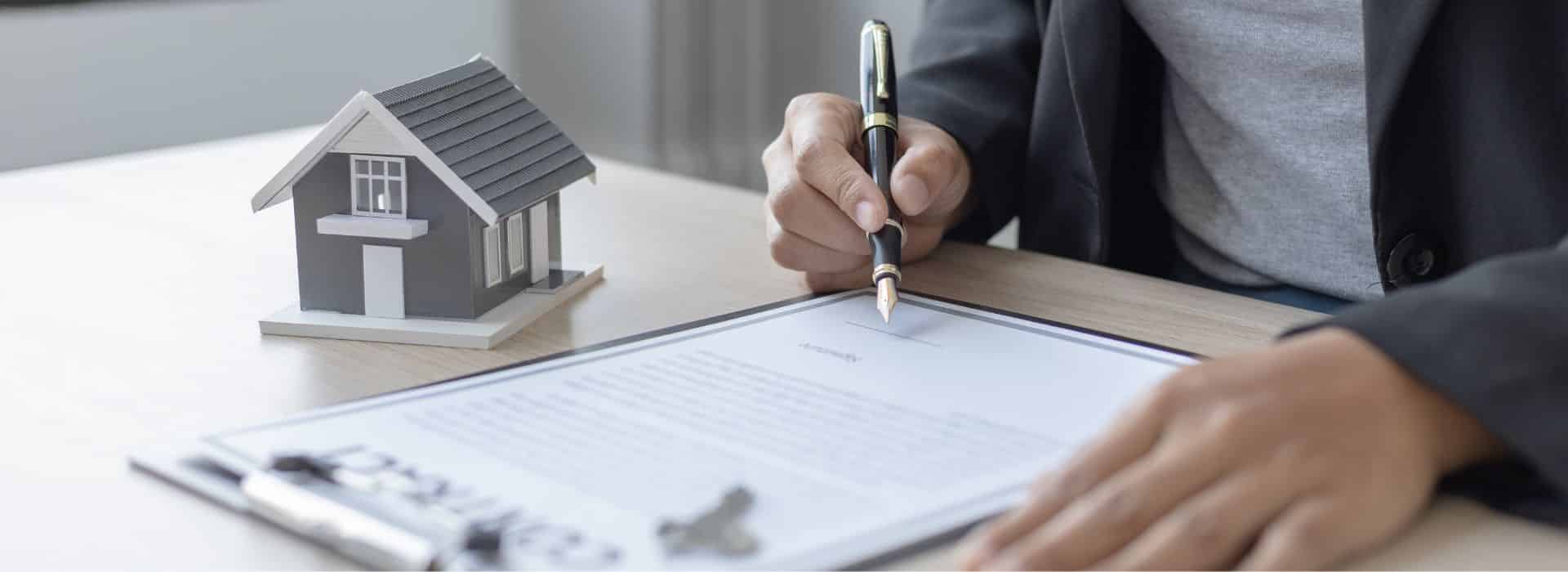 The image shows a person in business attire signing a document, with a small model house on the table, suggesting a real estate transaction.