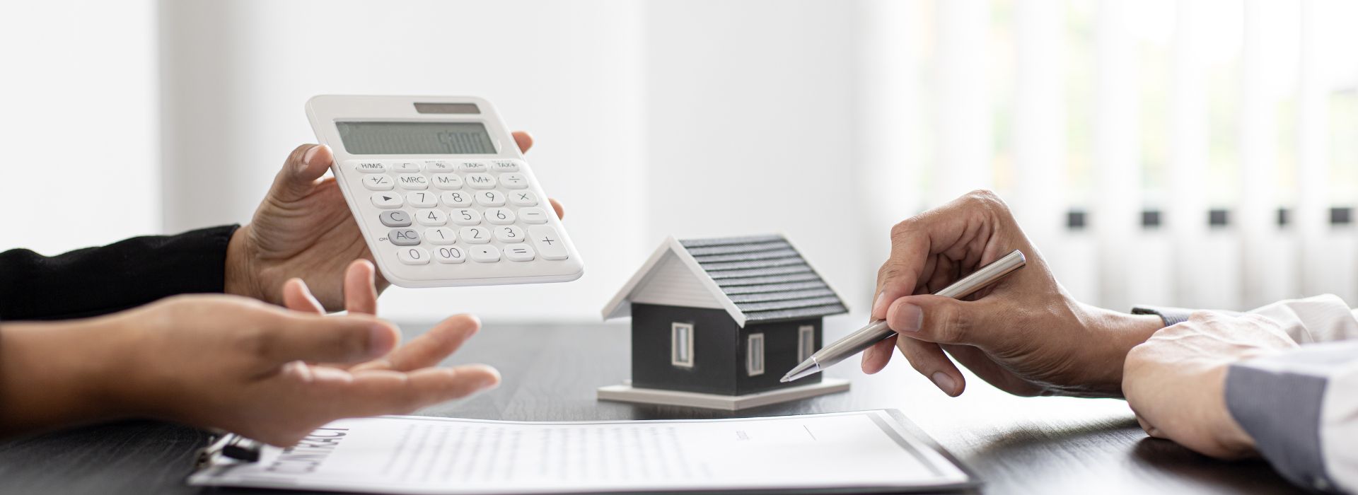 Two people at a white desk discussing finances with a calculator, small black house model, and paperwork. One person in black holds a white calculator while the other writes with a pen on documents, suggesting a real estate or mortgage consultation meeting.