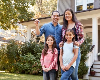 A happy family of four stands in front of their new home - parents and two young daughters posing together while the father holds up house keys. The house features gray siding, white trim, a covered porch, and is surrounded by mature trees and landscaping