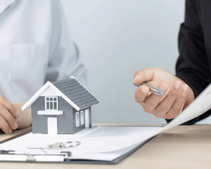 Two individuals sit at a desk discussing documents, with one person holding a pen and pointing to a paper. A small model house sits on the table between them, symbolizing a conversation about real estate, home loans, or mortgage agreements. A set of keys and a clipboard are also visible on the table.