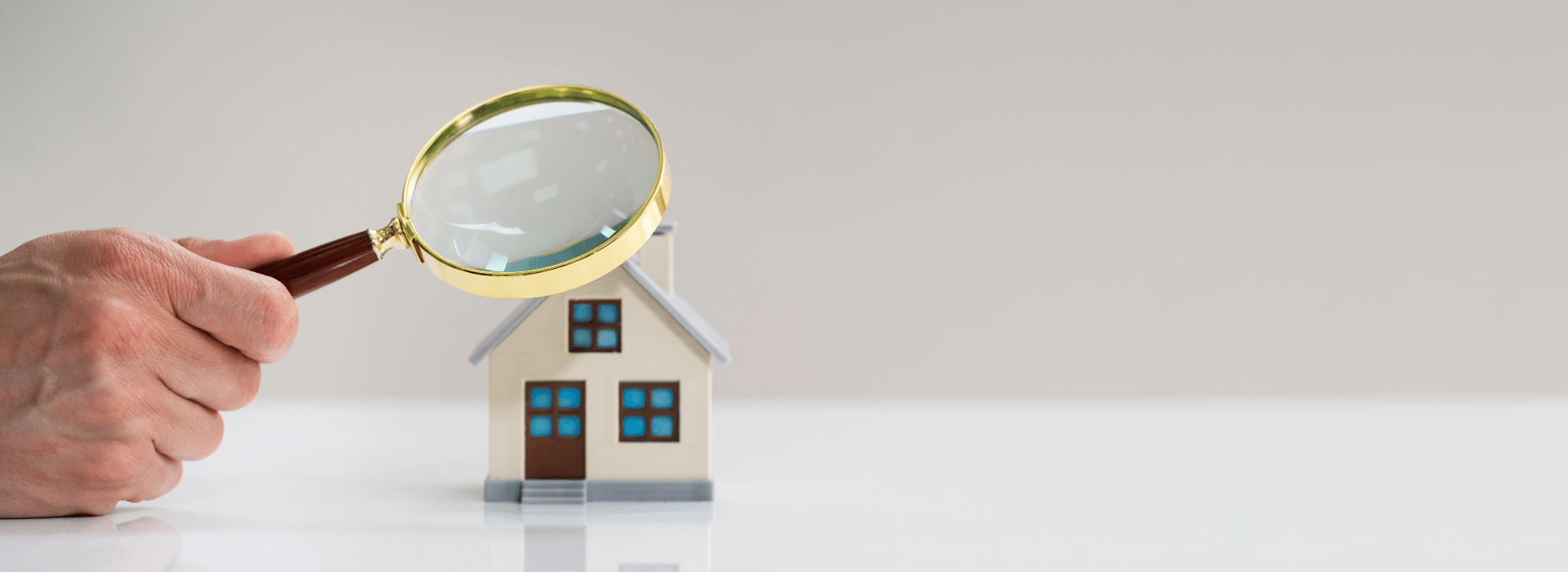 A hand holding a magnifying glass over a small white model house with blue windows and a brown door, symbolizing home inspection, property evaluation, or real estate search and analysis.