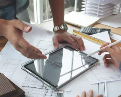 An architectural designer discusses home renovation on a tablet with a client at a desk, surrounded by building plans and a model of a house.