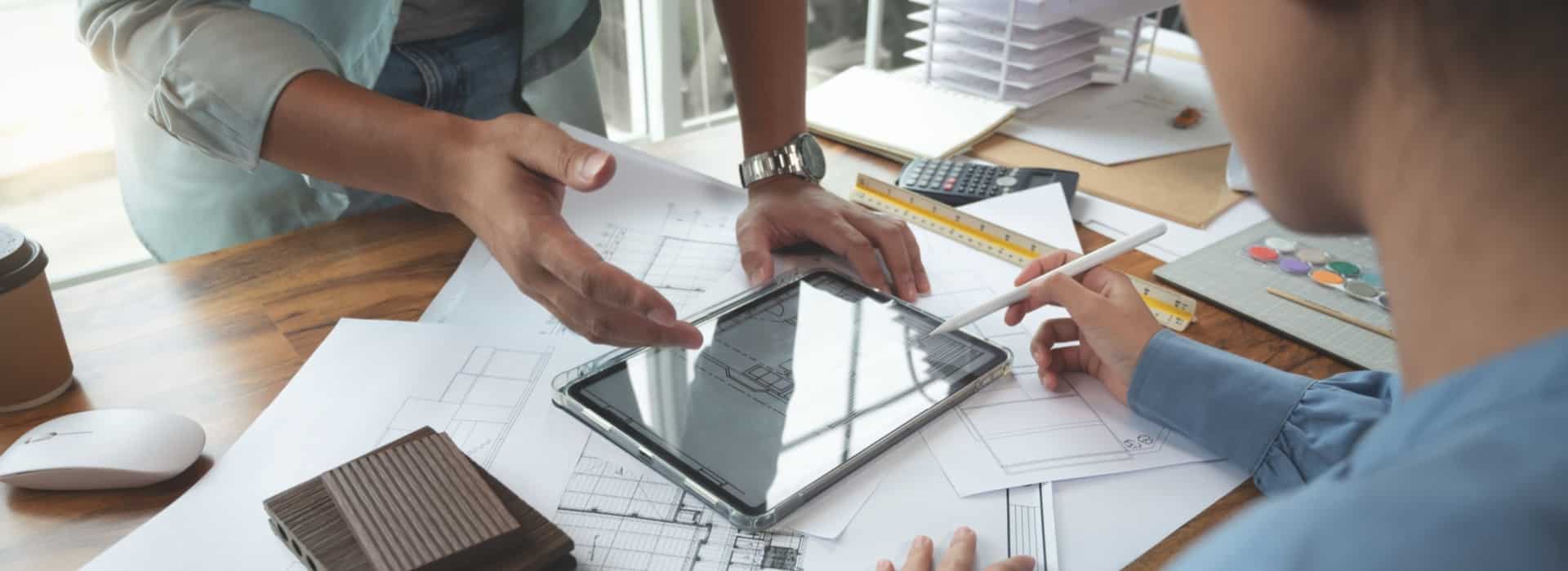 An architectural designer discusses home renovation on a tablet with a client at a desk, surrounded by building plans and a model of a house.